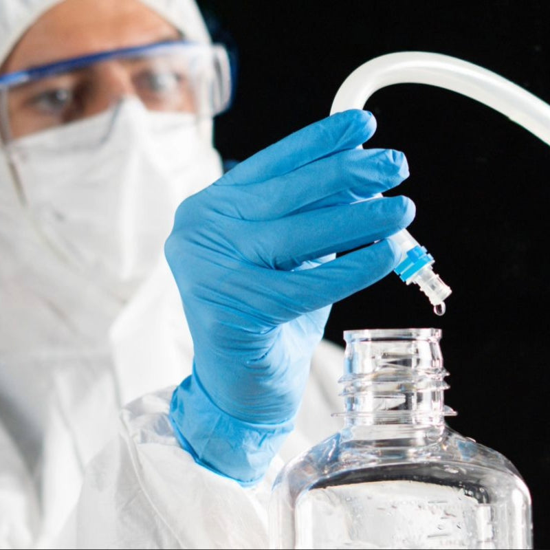 Operator in protective gear with blue gloves filling a clear single-use bottle with ADC. One drop misses the bottle.