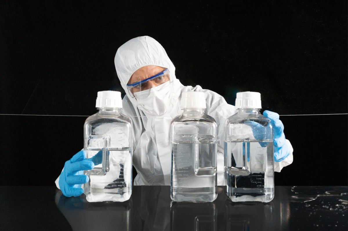 Person in protective gear handling large bottles of liquid in a laboratory setting. Bottles are not equally filled after manual handling