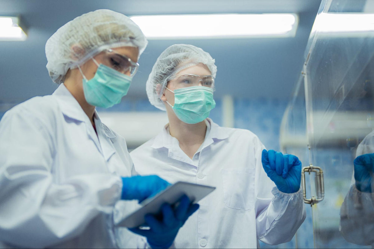 Two scientists in a sterile manufacturing environment, wearing protective clothing and discussing data on a tablet.