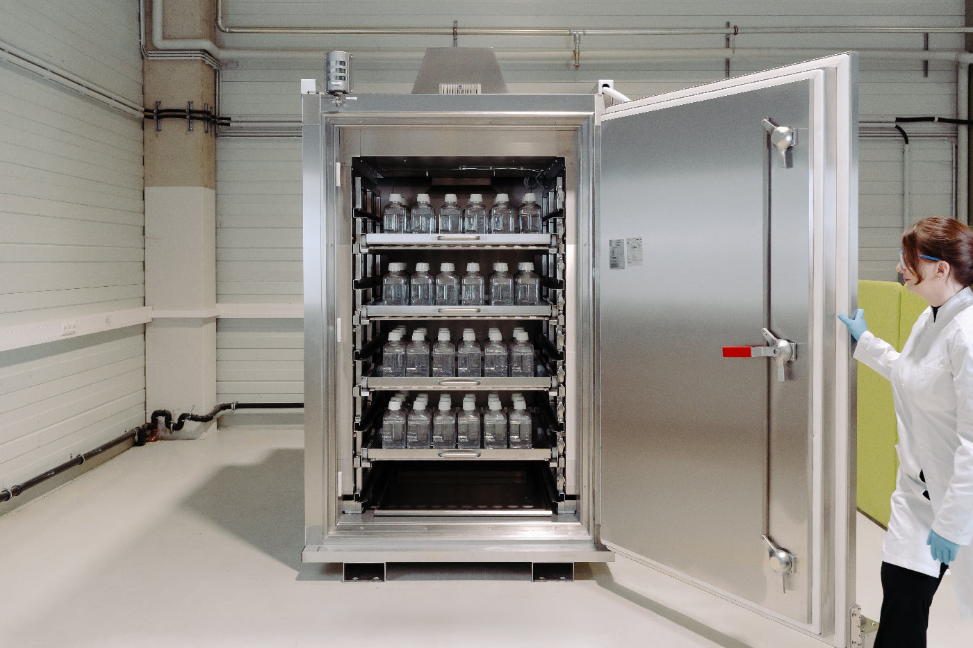 Person in a lab coat standing next to a large metal storage unit with shelves filled with bottles in a laboratory setting.